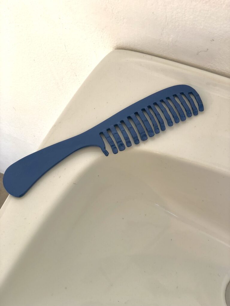 Steel sculpture of a big blue pocket comb with human fingers instead of teeth, resting on a bathroom sink.