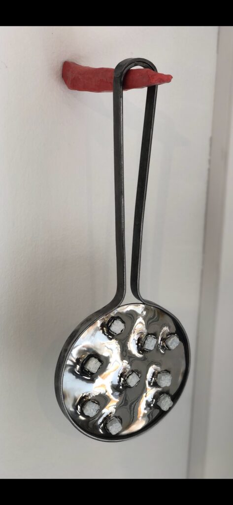 A steel sculpture of a hand mirror with protruding teeth hanging from a hook that looks like a tongue.