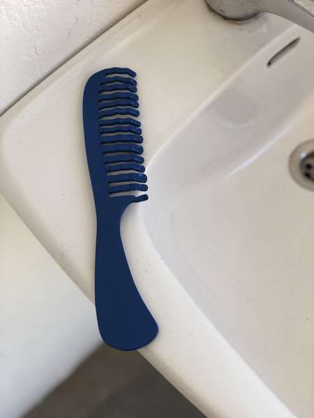 Steel sculpture of a big blue pocket comb with human fingers instead of teeth, resting on a bathroom sink.